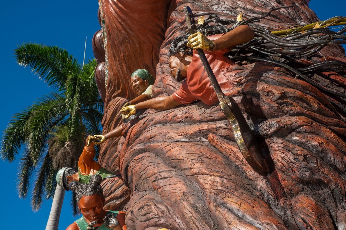The musician reaching across the carved bark toward the elder in a green head wrap