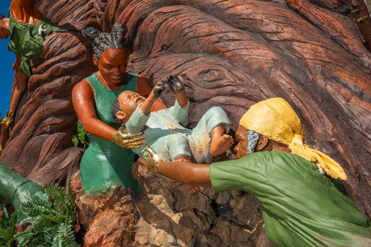 An elder in a yellow head wrap passing a crying baby up to a younger woman