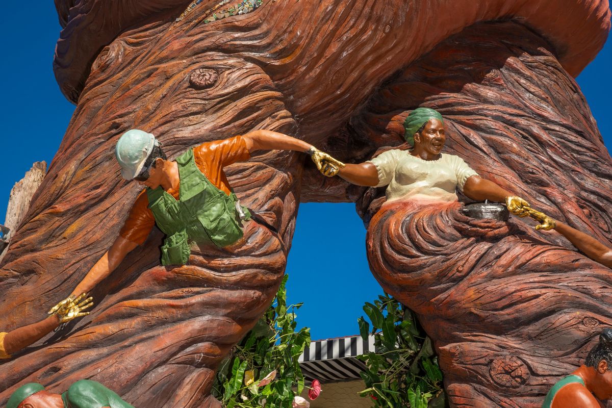 The builder in a hard hat and the elder in a green head wrap joining gilded hands across the arch
